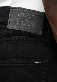 Μαύρο ύφασμα denim με δερμάτινη ετικέτα που φέρει ανεπίχριστη τη φράση "TOMMY JEANS" σε μπλε χρώμα. Τονίζεται από ένα μικρό κεντημένο λογότυπο στην τσέπη.