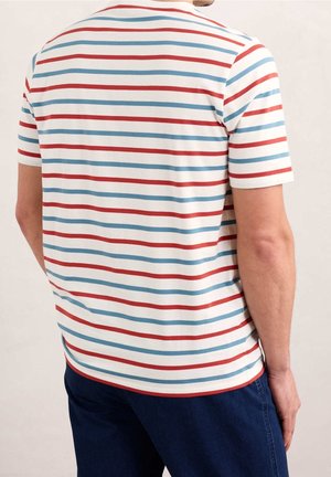 Uomo che indossa una camicia bianca a maniche corte con strisce orizzontali rosse e blu, abbinata a pantaloni blu scuro, in piedi con la schiena rivolta verso la fotocamera.