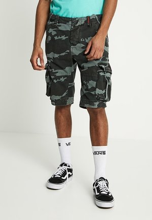 Man draagt zwarte en grijze camouflage cargoshorts, witte Vans sportsokken en zwarte Vans sneakers met witte veters tegen een effen achtergrond.