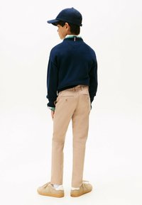 Ragazzo che indossa un maglione blu navy, pantaloni beige, sneakers beige, una camicia a quadri verde e un cappellino blu navy, in piedi e che guarda di lato.