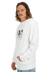 Biała bluza z kapturem z przednią kieszenią typu kangur, czarnym logo DC na piersi, sznurkiem do regulacji kaptura, ściągaczami przy mankietach i teksturowanym materiałem.