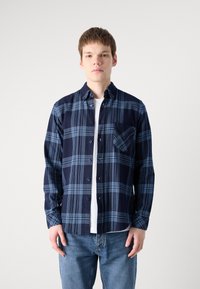 Camisa azul marinho e xadrez, de botões, mangas longas, um bolso no peito, tecido de algodão, usada sobre uma t-shirt branca, combinada com jeans azuis.