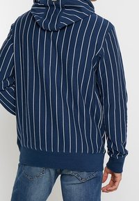 Marinblå hoodie med vita vertikala ränder, har en dragsko i huvan, ribbade ärmslut och nederkant samt en avslappnad passform. Blandning av bomull, mjuk textur.