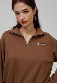 Rjav zip-up zgornji del s visokim ovratnikom, ki ima majhen zadrgo žep na prsih. Gladko blago z ohlapnim krojem in dolgimi rokavi.