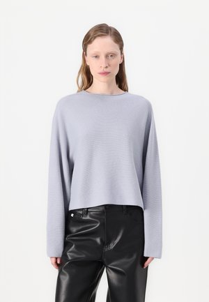 Hellgrauer, taillierter Pullover mit langen Ärmeln, hergestellt aus einem weichen Material. Kombiniert mit hoch taillierten schwarzen Lederhosen.
