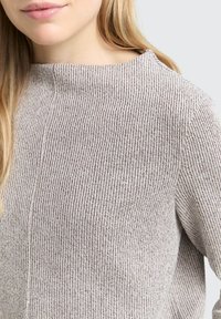 Jonge vrouw in een beige ribgebreide trui met lange mouwen en een bateau-uitsnijding, gedeeltelijk zichtbaar van de nek tot het midden van de torso.