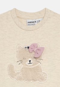 Beiges T-Shirt mit einem gestickten Katzendesign, das eine flauschige Textur und eine rosa Schleife auf dem Kopf der Katze zeigt. Rundhalsausschnitt, weicher Stoff.