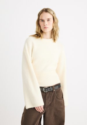 Žena s blond vlnitými vlasmi, oblečená v krémovom svetri s dlhým rukávom a hnedých nohaviciach s čiernym opaskom s cvočkami stojaca pred bielym pozadím.