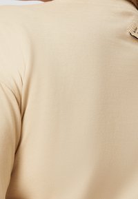 Nahaufnahme eines beigen T-Shirts aus strukturiertem Stoff mit einem kleinen gestickten Etikett mit der Aufschrift "Be your" in der Nähe des Ausschnitts.