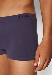 SLOGGI dunkelviolette Boxershorts mit glatter Oberfläche, ausgestattet mit einem weichen Bund und minimalen Nähtelementen. Die Hand des Modells liegt auf der Hüfte.