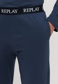 Pantaloni sport de culoare albastru închis dintr-un material moale, cu talie elastică și text alb "REPLAY". O mână este afișată în buzunarul lateral.