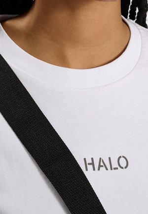 Camiseta de algodón blanca con cuello redondo y la impresión "HALO" en gris. Una tira negra es visible a través del hombro.