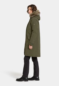 Oliven grøn parka med futteret hætte, sidelommer og lige snit, båret over mørke jeans og sorte ankelstøvler.