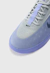 Grijze en paarse sport sneaker met een mesh bovenwerk, een gladde paarse rubberen zool en een contrasterend zwart logo. Voorzien van veters en een gestructureerd oppervlak.