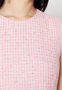 Vaaleanpunainen teksturoitu kangas, jossa on kudottu kuvio ja metalliset kultakorostukset. Pyöreä pääntie ja lyhyet hihat.