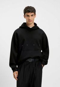 Black hooded sweatshirt with a relaxed fit, featuring a front kangaroo pocket and minimalistic white logo text across the chest. Smooth fabric texture.