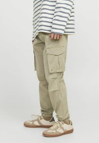Beige Cargo-Hosen mit mehreren Taschen, elastischen Bündchen und lässigem Schnitt, kombiniert mit cremefarbenen und braunen Sneakers sowie einem gestreiften Langarmhemd.