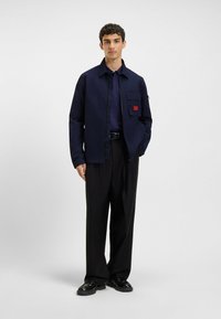 Navy zip-up jacket with chest pocket and red label detail, worn over a dark polo shirt and black pleated trousers, paired with black shoes.