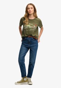 Olijfgroene graphic t-shirt met witte en rode tekst, gecombineerd met hoge blauwe denim jeans en groene high-top sneakers.