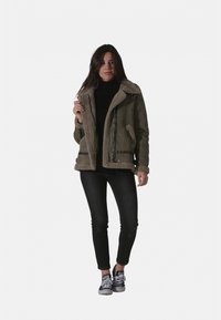 Veste vert olive avec un col et une doublure en shearling beige pelucheux, dotée de poches zippées, associée à un jean skinny noir et des baskets.