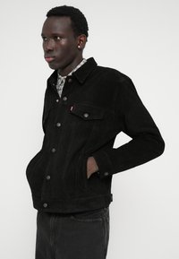 Μαύρη suedette τζάκετ τύπου trucker με γιακά, μπροστινές τσέπες, ασημί κουμπιά και αντίθετη ραφή, φορεμένη πάνω από ένα καρό πουκάμισο.
