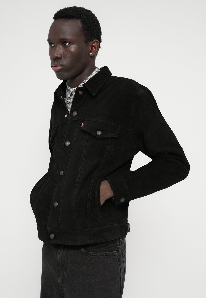 Giacca trucker in suede nera con colletto, tasche frontali, bottoni argentati e cuciture a contrasto, indossata sopra una camicia a quadri.