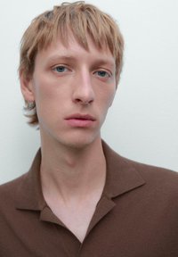 Close-up van een persoon met kort, lichtbruin haar, die een bruine polo met een zachte textuur draagt, tegen een lichtgrijze achtergrond.