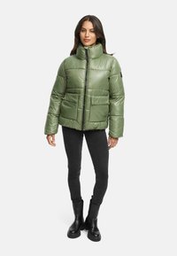 Grøn polstret jakke med høj krave, to frontlommer, lynlås lukning og en skinnende, glat tekstur. Båret over sorte leggings og støvler.