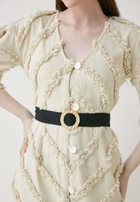 Beige mekko, jossa on tekstuurilla varustettuja sakara- ja syvään V-aukko. Siinä on valkoisia nappeja ja musta vyö kudotulla pyöreällä soljella.