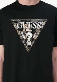 Svart bomull T-skjorte med et trekantet grafikk med et slitt gull- og hvitt "GUESS"-logo og spørsmålstegn-design.