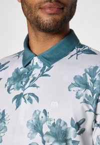 Kortermet polo skjorte i hvit med teal blomsterdesign, med teal krage, knappeåpning og brodert logo på brystet.