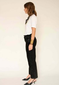 Jean taille haute en denim noir associés à un t-shirt blanc. Le modèle porte des chaussures à talons noires et des bracelets en or, mettant en valeur une silhouette élégante.