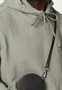 Sudadera gris con capucha, que incluye un cordón ajustable. Adjunto hay una bolsa circular de cuero negro con una correa y herrajes metálicos.