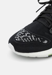 Czarny but sportowy z teksturowaną górą z dzianiny i białą podeszwą. Posiada gładki czarny panel pięty oraz odblaskowe detale na palcach.