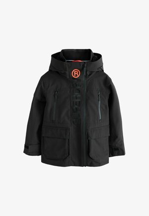 Barn, der står og bærer en sort hættetrøje, hvid skjorte, lyseblå jeans og sorte og hvide sneakers mod en ensfarvet baggrund.