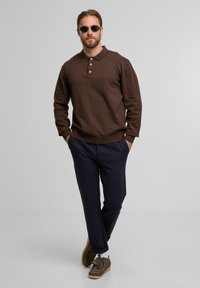 Mann in braunem Langarm-Polohemd, blauen Hosen, braunen Schuhen und Sonnenbrille, der mit den Händen in den Taschen vor einem schlichten hellgrauen Hintergrund läuft.