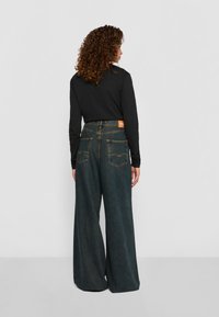 Persoon met krullend haar, gekleed in een zwart langslavend shirt en high-waisted, wijde donkerblauwe jeans, staande tegen een effen achtergrond.