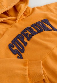 Sudadera naranja con un gran gráfico bordado en azul marino de "SUPERDRY" en la parte frontal. Presenta un bolsillo tipo canguro y puños de canalé. Textura suave.