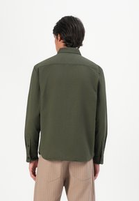 Camiseta de manga larga verde oliva con cuello, puños abotonados y dobladillo recto, combinada con pantalones rayados ligeros beige.