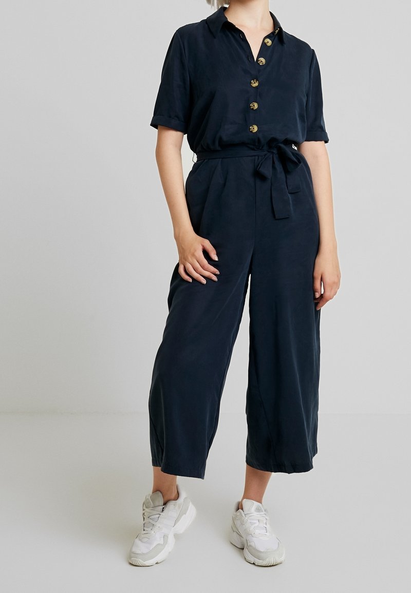 Femme portant une combinaison bleu marine à boutons, manches courtes, ceinture à la taille, jambes larges raccourcies et baskets blanches épaisses sur un fond uni.