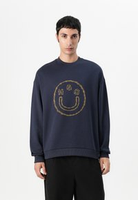 Sudadera marinera con un logotipo redondo bordado en dorado que presenta "HGO" y un diseño de carita sonriente. Fabricada en un suave tejido texturizado con cuello redondo.
