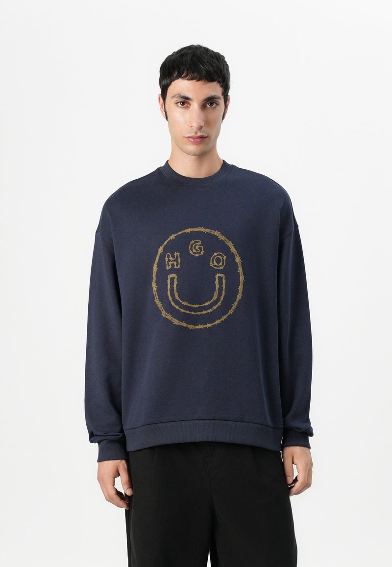 Sudadera marinera con un logotipo redondo bordado en dorado que presenta "HGO" y un diseño de carita sonriente. Fabricada en un suave tejido texturizado con cuello redondo.