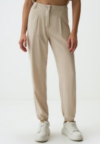 Pantaloni beige a pieghe con chiusura a bottoni, polsini elasticizzati e una texture liscia, abbinati a sneaker bianche su uno sfondo neutro.
