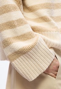 Μανίκι πλεκτού πουλόβερ με ρίγες σε μπεζ και εκρού σε χέρι ατόμου με το χέρι μέσα στην τσέπη μπεζ παντελονιού.