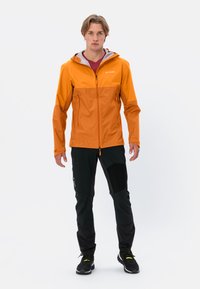 Giacca impermeabile arancione con dettagli grigi, zip frontale e due tasche sul petto. Abbinata a pantaloni neri con pannelli testurizzati.