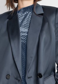 Blazer azul marino de satén con botones negros, combinado con una camisa azul texturizada con un patrón de diamantes. Corte entallado con solapas afiladas.
