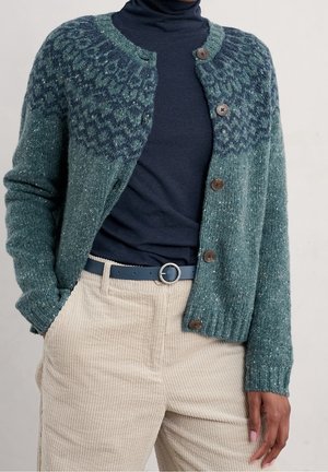 Osoba ubrana w niebieski wzorzysty sweter kardigan na guziki, pod którym ma granatowy golf, beżowe spodnie sztruksowe oraz niebieski pasek z okrągłą srebrną klamrą.