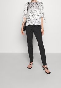 Vrouw in een zilveren blouse met stippen en geknoopte mouwen, zwarte slim-fit broeken, zwarte gespsandalen met hakken en een zwarte schoudertas.