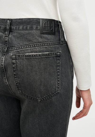 Close-up of a person wearing dark gray denim jeans with a black logo patch on the waistband and a white long-sleeve top.