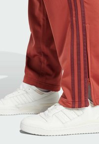 Pantalons de sport rouges avec des poignets zippés, ornés de trois bandes rouge foncé sur les côtés, associés à des baskets blanches.